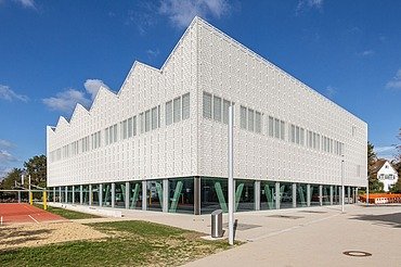 Außenansicht einer modernen Sporthalle mit heller Ziegelfassade, rhythmisch angeordneten Oberlichtern und grünen Stahlstützen im Erdgeschoss. Im Vordergrund befinden sich Sportflächen und Wege, umgeben von Bäumen und Schulgebäuden.