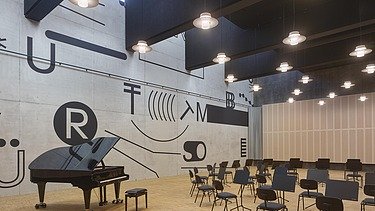 Innenansicht eines modernen Probenraums mit großer grafisch gestalteter Sichtbetonwand, mehreren Stühlen und einem Flügel auf Holzparkett. Über dem Raum hängen akustisch optimierte Leuchten und Deckenpaneele, die den Saal für Musikproben oder Aufführungen ausstatten.