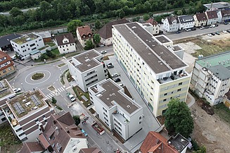 Luftaufnahme eines Wohngebäudekomplexes mit mehreren mehrstöckigen Häusern, angrenzenden Straßen und Grünflächen im Hintergrund.