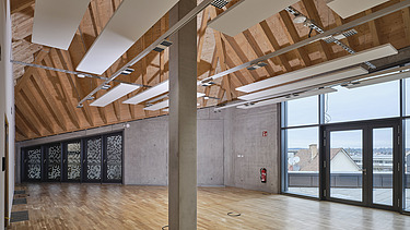 Innenansicht eines großzügigen, modernen Raumes mit schräger Holzdecke, akustischen Deckenpaneelen, großen Glasfronten und Parkettboden. Der Raum wirkt lichtdurchflutet und bietet einen Blick auf die Stadt.