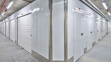 Gang mit mehreren weißen Selfstorage-Einheiten, ausgestattet mit Rolltoren und Metallverkleidungen. Sichtbare Betonboden, Decke mit Rohrleitungen und Beleuchtung.