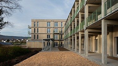 Außenansicht eines neu errichteten Wohngebäudes mit vertikaler Holzfassade, Balkonen und einem durchgehenden Laubengang aus Betonstützen und grünen Geländern. Im Vordergrund liegt eine frisch angelegte Außenfläche, im Hintergrund sind Wohnhäuser und Landschaft erkennbar. Moderne, energieeffiziente Wohnarchitektur.