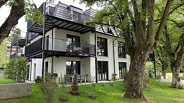 Mehrfamilienhäuser mit dunklen Mansarddächern, großen Balkonen und grüner Gartenanlage unter blauem Himmel mit Wolken.
