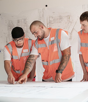 Drei Personen in orangefarbenen Sicherheitswesten stehen an einem Tisch und betrachten Baupläne, im Hintergrund hängen technische Zeichnungen an der Wand.