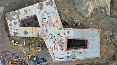Luftaufnahme einer Baustelle mit einem großen, 8-förmigen Gebäudefundament in der Rohbauphase. Auf der Betonplatte sind Baumaterialien, Baugeräte und Gerüste verteilt. Rund um die Baustelle befinden sich Sandflächen, Baumaschinen und Container.