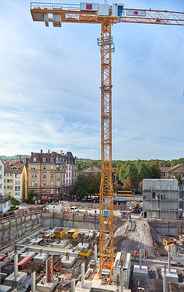 Große Baustelle mit gelbem Turmdrehkran, Betonfundamenten und Baugeräten, umgeben von mehrstöckigen Wohngebäuden und Bäumen unter blauem Himmel.