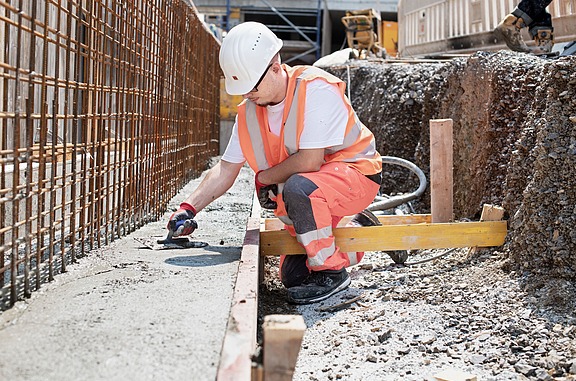 Azubi in Sicherheitskleidung glättet frischen Beton in einer Schalung auf einer Baustelle, umgeben von Bewehrungsstahl.