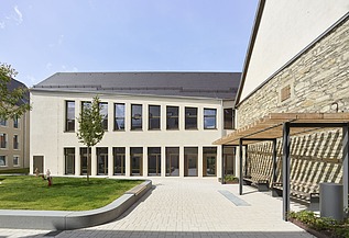 Innenhof eines modernen Gebäudekomplexes mit heller Putzfassade, großen Fensterfronten und einer gepflegten Grünfläche. Rechts befindet sich eine historische Natursteinwand mit Holzpergola, links ein kleiner Baum und Sitzbereiche. Der Hof ist offen gestaltet und mit hellen Pflastersteinen ausgelegt.