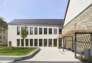Innenhof eines modernen Gebäudekomplexes mit heller Putzfassade, großen Fensterfronten und einer gepflegten Grünfläche. Rechts befindet sich eine historische Natursteinwand mit Holzpergola, links ein kleiner Baum und Sitzbereiche. Der Hof ist offen gestaltet und mit hellen Pflastersteinen ausgelegt.