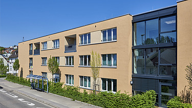 Ansicht eines dreigeschossigen Wohngebäudes mit sandfarbener Fassade und großen Fenstern. Rechts befindet sich ein markanter Glasbereich mit Eingangszone, davor eine begrünte Hecke entlang des Gehwegs. Die Straße im Vordergrund ist mit Bushaltestelle und Fahrbahnmarkierungen versehen, im Hintergrund blauer Himmel.