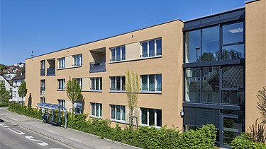 Ansicht eines dreigeschossigen Wohngebäudes mit sandfarbener Fassade und großen Fenstern. Rechts befindet sich ein markanter Glasbereich mit Eingangszone, davor eine begrünte Hecke entlang des Gehwegs. Die Straße im Vordergrund ist mit Bushaltestelle und Fahrbahnmarkierungen versehen, im Hintergrund blauer Himmel.
