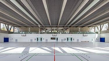 Innenansicht einer Dreifeldsporthalle mit hellem Sportboden und farbigen Markierungen. Die Decke besteht aus Sichtbetonträgern und akustischen Paneelen. Entlang der Stirnseite befinden sich Glasflächen mit Blick auf den umliegenden Gebäudetrakt.