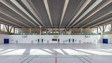 Innenansicht einer Dreifeldsporthalle mit hellem Sportboden und farbigen Markierungen. Die Decke besteht aus Sichtbetonträgern und akustischen Paneelen. Entlang der Stirnseite befinden sich Glasflächen mit Blick auf den umliegenden Gebäudetrakt.