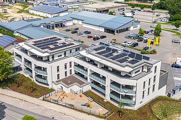 Luftaufnahme eines modernen Gebäudekomplexes mit heller Fassade und großen Balkonen. Auf den Flachdächern sind Solarpaneele installiert. Das Gebäude ist von Straßen, Grünflächen und weiteren Gewerbebauten umgeben.