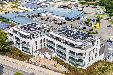 Luftaufnahme eines modernen Gebäudekomplexes mit heller Fassade und großen Balkonen. Auf den Flachdächern sind Solarpaneele installiert. Das Gebäude ist von Straßen, Grünflächen und weiteren Gewerbebauten umgeben.