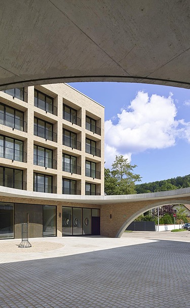 Blick in den Innenhof eines modernen Gebäudes mit Klinkerfassade, gesehen durch eine große ovale Dachöffnung aus Sichtbeton. Der Hof ist gepflastert, umliegende Gebäude haben regelmäßige Fensterreihen, und im Hintergrund sind Grünflächen sowie blauer Himmel mit Wolken sichtbar.