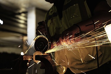 Nahaufnahme einer Person in Sicherheitsweste und Helm. Helle Funken sprühen vom Schleifpunkt in verschiedene Richtungen, während der Hintergrund dunkel und unscharf bleibt.