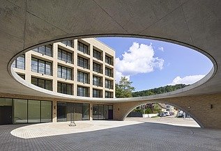 Blick in den Innenhof eines modernen Gebäudes mit Klinkerfassade, gesehen durch eine große ovale Dachöffnung aus Sichtbeton. Der Hof ist gepflastert, umliegende Gebäude haben regelmäßige Fensterreihen, und im Hintergrund sind Grünflächen sowie blauer Himmel mit Wolken sichtbar.