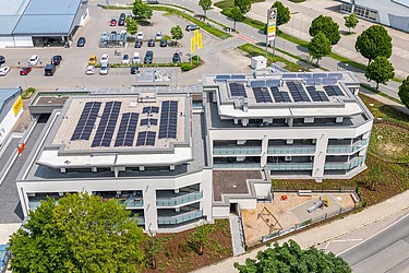 Luftaufnahme eines modernen Gebäudekomplexes mit großen Solarpanelen auf den Dächern, umgeben von Straßen, Grünflächen und angrenzenden Gewerbegebäuden.