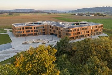 Luftaufnahme eines großen, modernen Gebäudes mit klarer Architektur und flachen Dächern, umgeben von weitläufigen Feldern und Grünflächen. Im Vordergrund sind Bäume sichtbar, im Hintergrund erstreckt sich eine hügelige Landschaft unter blauem Himmel.