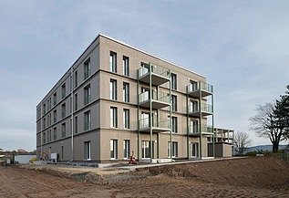 Außenansicht eines viergeschossigen Wohngebäudes mit klarer, moderner Fassadengestaltung und mehreren Balkonen. Das Gebäude steht noch auf einer unbefestigten Baustellenfläche, im Hintergrund Bäume und offener Himmel.