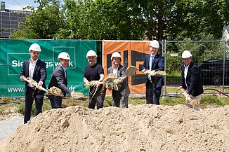 Baustelle mit Betongebäuden, Stahlträgern und Bauarbeitern mit Sicherheitsausrüstung unter teils bewölktem Himmel