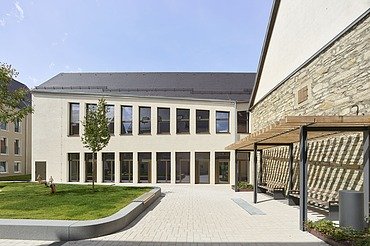 Innenhof eines modernen Gebäudekomplexes mit heller Putzfassade, großen Fensterfronten und einer gepflegten Grünfläche. Rechts befindet sich eine historische Natursteinwand mit Holzpergola, links ein kleiner Baum und Sitzbereiche. Der Hof ist offen gestaltet und mit hellen Pflastersteinen ausgelegt.