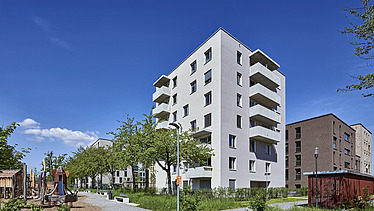 Mehrstöckiges Wohngebäude in heller Fassade mit Balkonen, umgeben von Grünflächen, Bäumen und einem gepflasterten Weg unter klarem blauem Himmel.