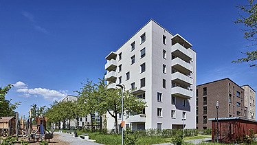 Mehrstöckiges Wohngebäude in heller Fassade mit Balkonen, umgeben von Grünflächen, Bäumen und einem gepflasterten Weg unter klarem blauem Himmel.