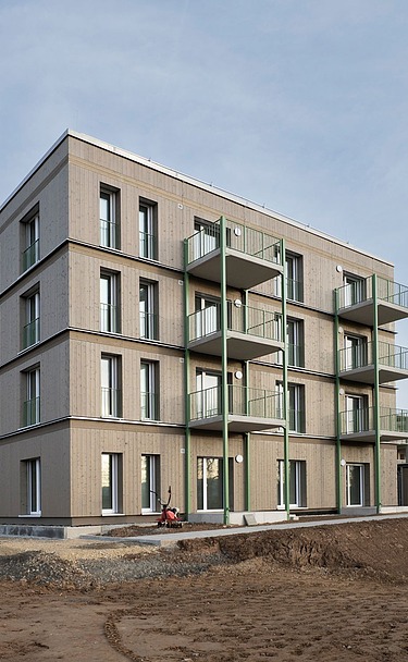 Außenansicht eines viergeschossigen Wohngebäudes mit klarer, moderner Fassadengestaltung und mehreren Balkonen. Das Gebäude steht noch auf einer unbefestigten Baustellenfläche, im Hintergrund Bäume und offener Himmel.