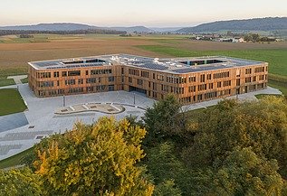 Luftaufnahme eines großen, modernen Gebäudes mit klarer Architektur und flachen Dächern, umgeben von weitläufigen Feldern und Grünflächen. Im Vordergrund sind Bäume sichtbar, im Hintergrund erstreckt sich eine hügelige Landschaft unter blauem Himmel.