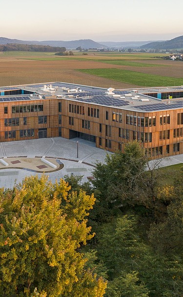 Luftaufnahme eines großen, modernen Gebäudes mit klarer Architektur und flachen Dächern, umgeben von weitläufigen Feldern und Grünflächen. Im Vordergrund sind Bäume sichtbar, im Hintergrund erstreckt sich eine hügelige Landschaft unter blauem Himmel.