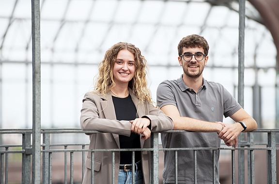 Zwei Studierende stehen an einem Metallgeländer in einer offenen, überdachten Struktur mit Stahlträgern und Glasdach.