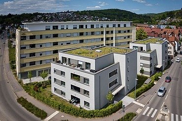 Luftaufnahme einer zeitgemäßen Wohnanlage mit mehreren Gebäuden, hellen Fassaden und begrünten Flachdächern. Die Anlage liegt an einer Straßenecke, umgeben von Gehwegen und Grünflächen, mit Blick auf eine hügelige Landschaft im Hintergrund.