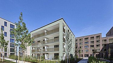 Mehrstöckiges Wohnhaus in moderner Architektur mit großen Balkonen, umgeben von gepflegten Grünflächen und jungen Bäumen, unter klarem blauem Himmel.