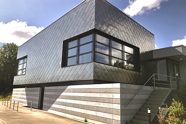 Zeitgemäßes Gebäude mit dunkler Metallfassade, großen Fenstern und seitlicher Treppe unter blauem Himmel