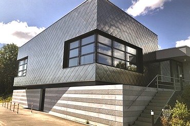 Zeitgemäßes Gebäude mit dunkler Metallfassade, großen Fenstern und seitlicher Treppe unter blauem Himmel