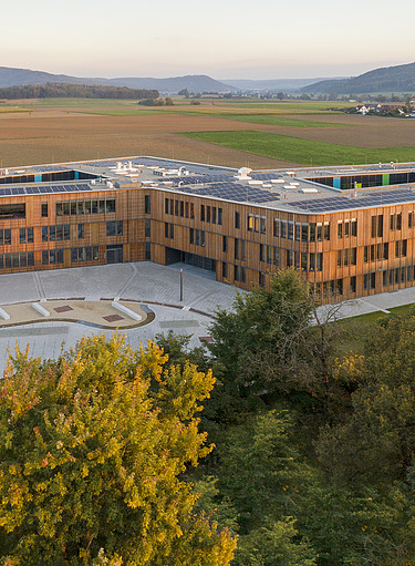 Moderne, mehrstöckige Realschule mit Flachdach und großen Fensterfronten in einer grünen Landschaft.