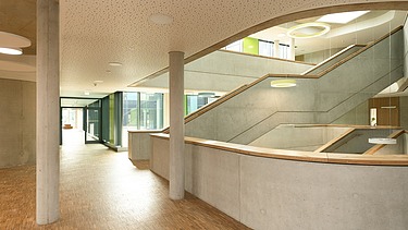Innenansicht eines großzügigen Gebäudes mit Sichtbetonwänden, breiter Treppe und geschwungenem Geländer. Der Boden ist aus Holz, die Decke mit akustischen Elementen versehen. Große Glasflächen im Hintergrund sorgen für viel Tageslicht und eine helle Atmosphäre.