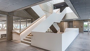 Baustelle mit Betongebäuden, Stahlträgern und Bauarbeitern mit Sicherheitsausrüstung unter teils bewölktem Himmel