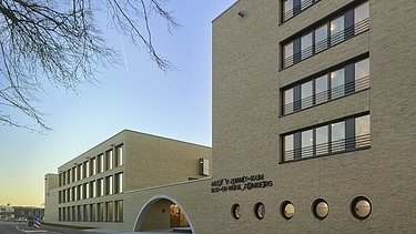 Außenansicht eines modernen Gebäudekomplexes mit heller Klinkerfassade, großen Fensterbändern und einem prägnanten Rundbogen-Eingang. Die Architektur kombiniert klare Linien mit kreisförmigen Fensteröffnungen entlang der Straße, während der Abendhimmel eine warme Lichtstimmung erzeugt.