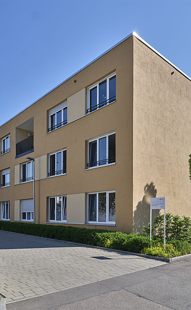 Ansicht eines modernen, dreigeschossigen Wohngebäudes mit hellgelber Fassade, großen Fenstern und Balkonen. Vor dem Gebäude befinden sich gepflegte Grünflächen und eine gepflasterte Zufahrt, im Hintergrund sind Bäume und blauer Himmel sichtbar.