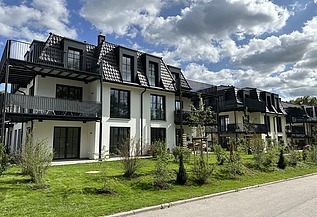 Mehrfamilienhäuser mit dunklen Dächern, großen Balkonen und grüner Gartenanlage unter blauem Himmel mit Wolken.
