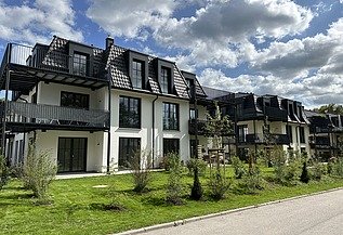 Mehrfamilienhäuser mit dunklen Dächern, großen Balkonen und grüner Gartenanlage unter blauem Himmel mit Wolken.