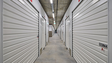 Langer Gang mit weißen Selfstorage-Einheiten, ausgestattet mit stabilen Rolltoren und Metallgriffen. Betonboden und Decke mit sichtbarer Beleuchtung und Rohrleitungen sorgen für eine funktionale, sichere Lagerumgebung.