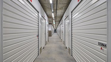 Langer Gang mit weißen Selfstorage-Einheiten, ausgestattet mit stabilen Rolltoren und Metallgriffen. Betonboden und Decke mit sichtbarer Beleuchtung und Rohrleitungen sorgen für eine funktionale, sichere Lagerumgebung.