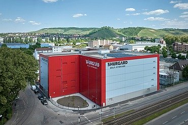 Großes Selfstorage-Gebäude mit roter und weißer Fassade, gelegen an einer Straße mit Straßenbahnschienen. Im Hintergrund grüne Hügel, Wohnhäuser und blauer Himmel mit wenigen Wolken.