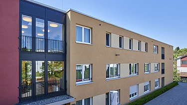 Ansicht eines zeitgemäßen Wohngebäudes mit klarer Linienführung, hellgelber Fassade und einem markanten dunklen Glasbereich mit Balkon. Die Architektur kombiniert große Fensterflächen mit schlichten Formen, im Hintergrund blauer Himmel und Grünflächen.