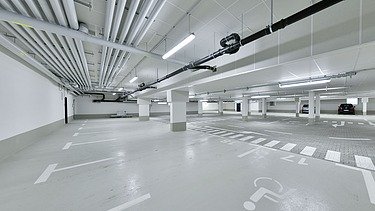 Leere Tiefgarage mit weißen Stellplatzmarkierungen, breiten Fahrwegen und Deckenrohren.