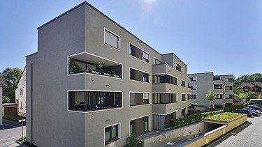 Ansicht eines modernen Mehrfamilienhauses mit klaren Linien, großen Balkonen und hellgrauer Fassade. Im Vordergrund befindet sich eine Zufahrt zur Tiefgarage, umgeben von Grünflächen. Der Himmel ist blau und sonnig, was die helle Architektur betont.